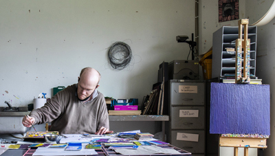 Tom O'Sullivan sits at a desk with colourful papers. He dips his paintbrush into paint. Behind him are arts materials and a filing cabinet. To his left is a canvas painted in blue on an easel