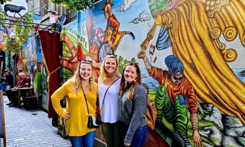 Three women smiling in front of a mural near the English Market
