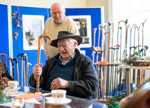 Man wearing a black hat sits at a table laughing, holding a cane in his right hand