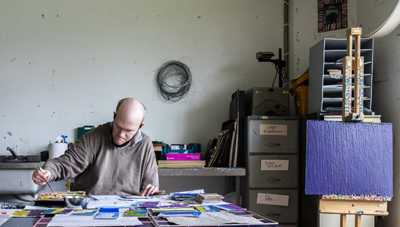 Artist sits at their desk in their studio, there are colourful pages spread out on the table, an easel stands to the right of the desk