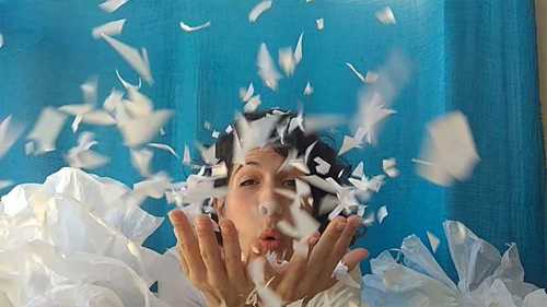 Person blows small pieces of white paper towards camera