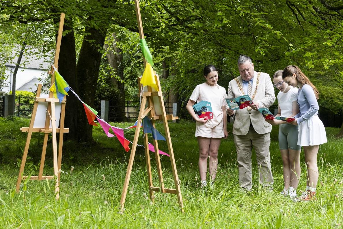 Lord Mayor Cllr Dan Boyle stands with three young girls in long grass, looking at the Cruinniu na nÓg 2025 programme. Two easels are on the left of the image with bunting hanging between therm.